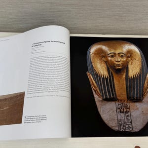 May include: A book open to a page with text and an image of a golden mask. The mask has a serene face with gold and blue details. The book page includes text about the mask and a smaller image of a brown surface.