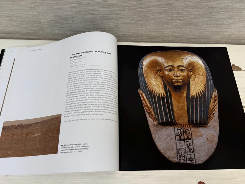 May include: A book open to a page with text and an image of a golden mask. The mask has a serene face with gold and blue details. The book page includes text about the mask and a smaller image of a brown surface.