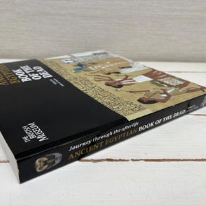 May include: A hardcover book titled "ANCIENT EGYPTIAN BOOK OF THE DEAD" from The British Museum. The book has a black spine and a cover featuring ancient Egyptian artwork. The book's title and author are visible.
