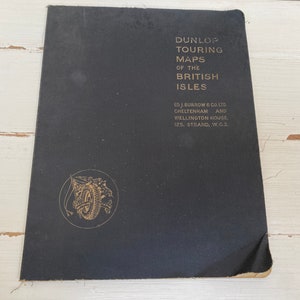 May include: Black cover of a book titled "Dunlop Touring Maps of the British Isles" with a gold logo of a tire and a motorcycle. The book is published by Ed. J. Burrow & Co. Ltd. Cheltenham and Wellington House, 125. Strand, W.G.2.