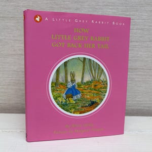 以下が含まれることがあります： 「How Little Grey Rabbit Got Back Her Tail」という金色の文字が書かれたピンク色の本。表紙には、青いドレスを着たウサギが森の中を歩いているイラストが描かれている。
