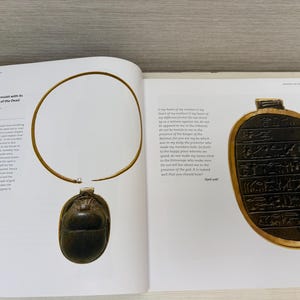 May include: An open book displays a gold-colored necklace with a scarab pendant and an oval-shaped amulet with hieroglyphs. The necklace has a rigid neck band. The pendant is dark brown and beetle-shaped.