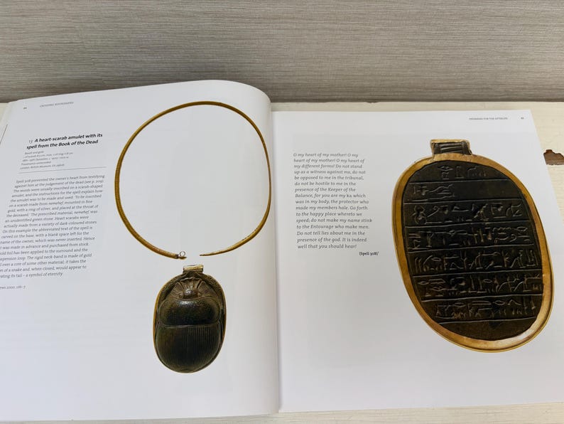 May include: An open book displays a gold-colored necklace with a scarab pendant and an oval-shaped amulet with hieroglyphs. The necklace has a rigid neck band. The pendant is dark brown and beetle-shaped.