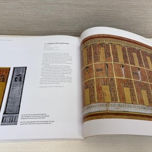 May include: An open book displaying pages with ancient Egyptian art and hieroglyphs. One page features colorful rectangular panels with inscriptions. The opposite page shows a detailed illustration of a wall with figures and more hieroglyphs. The book is open on a white surface.