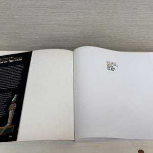 May include: An open book titled "Ancient Egyptian Book of the Dead." The left page displays a black cover with text and an illustration of an Egyptian statue. The right page is blank with the title printed at the top.