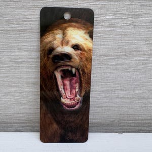 May include: A rectangular bookmark featuring a detailed illustration of a brown bear with its mouth wide open, revealing teeth and a pink interior. The bookmark has a hole at the top.