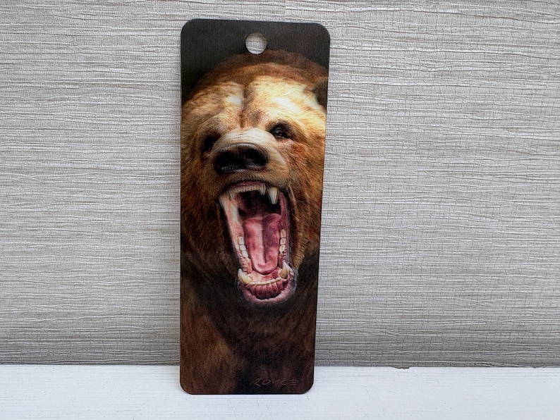 May include: A rectangular bookmark featuring a detailed illustration of a brown bear with its mouth wide open, revealing teeth and a pink interior. The bookmark has a hole at the top.