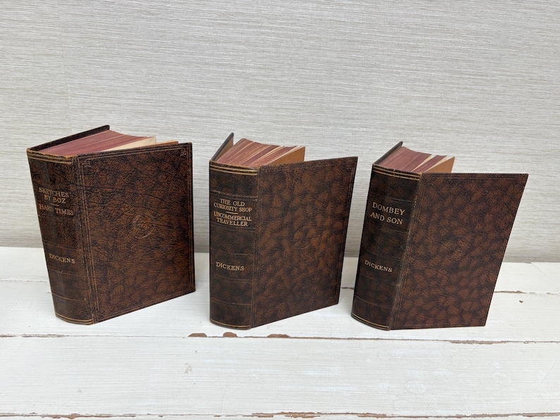 May include: Three brown decorative book-shaped boxes with titles like "Sketches by Boz" and "Dombey and Son" by Dickens. The boxes have a textured leather-like appearance and red-toned pages. The books are arranged on a white surface.