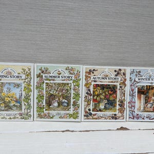 Puede incluir: Un conjunto de cuatro libros de Brambly Hedge por Jill Barklem titulados Spring Story, Summer Story, Autumn Story y Winter Story. Cada portada presenta una ilustración detallada y colorida de ratones en entornos estacionales.
