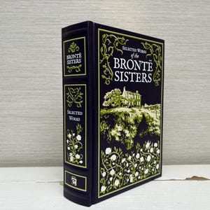 Puede incluir: Un libro de tapa dura titulado "Selected Works of the Brontë Sisters". El libro tiene una cubierta negra con detalles decorativos dorados y verdes. El lomo presenta el texto "Brontë Sisters" y "Selected Works".
