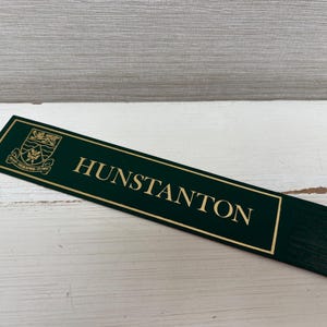 May include: A dark green rectangular sign with gold lettering that reads "HUNSTANTON". The sign has a gold border and a crest in the upper left corner. The sign is on a white wooden surface.