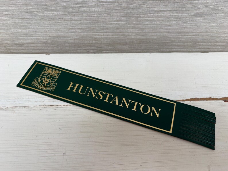 May include: A dark green rectangular sign with gold lettering that reads "HUNSTANTON". The sign has a gold border and a crest in the upper left corner. The sign is on a white wooden surface.