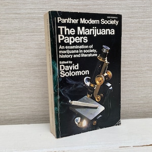 Può includere: Un libro in bianco e nero intitolato "The Marijuana Papers" con un microscopio sulla copertina. Il libro tratta della marijuana nella società, nella storia e nella letteratura. È curato da David Solomon.