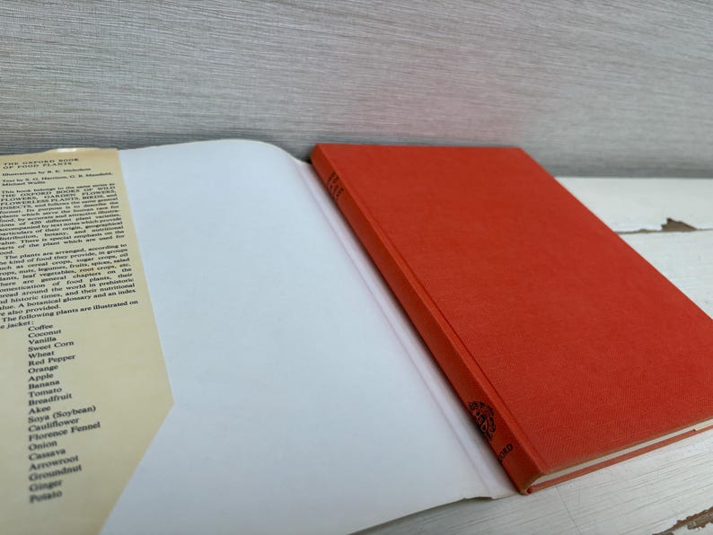 May include: An open book with a red cover and white pages. The left page has text listing plants such as coffee, vanilla, and tomato. The book is on a white surface.