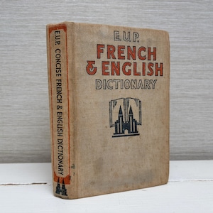 Peut inclure: Un livre vintage brun avec le titre "E.U.P. French & English Dictionary" imprimé en lettres noires et rouges. La couverture du livre présente une illustration en noir et blanc d'un bâtiment avec deux tours.