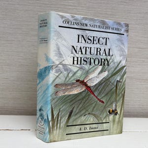 Puede incluir: Un libro de tapa dura titulado "Insect Natural History" de la serie Collins New Naturalist. La portada presenta una ilustración detallada de una libélula y una abeja entre hierbas altas. El lomo del libro muestra el título y el nombre del autor.