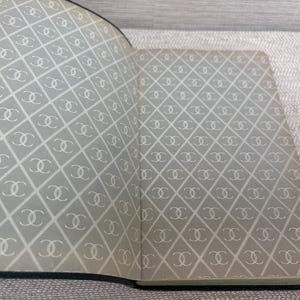 May include: Open notebook with a gray and white diamond pattern featuring interlocking double C logos. The notebook has a dark green border and is open to reveal the patterned interior pages.