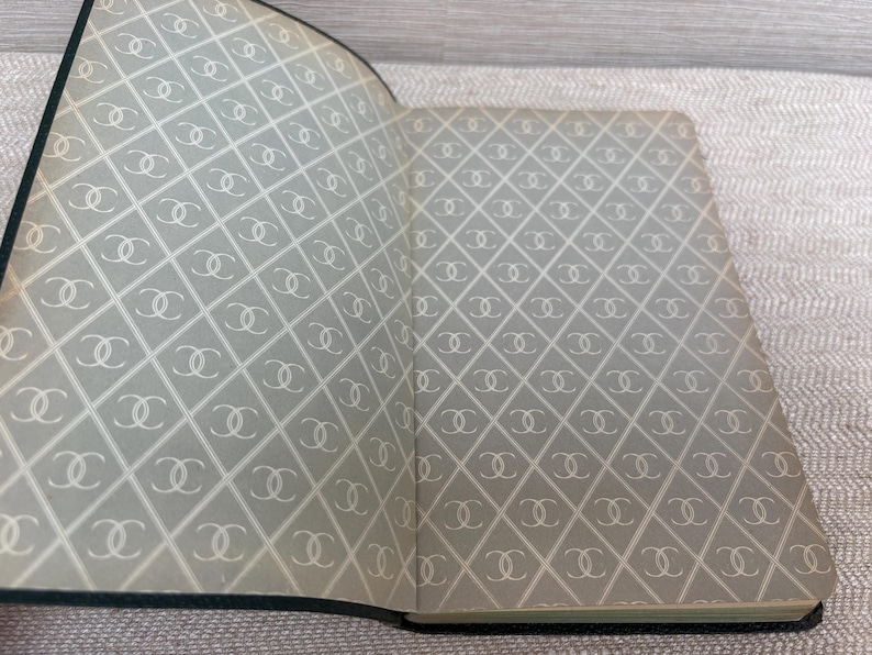 May include: Open notebook with a gray and white diamond pattern featuring interlocking double C logos. The notebook has a dark green border and is open to reveal the patterned interior pages.