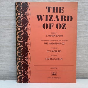 Puede incluir: Una portada de libro rosa con un borde negro con un diseño floral. El título es "El mago de Oz" con el autor listado como "L Frank Baum". El libro es un libreto para el musical basado en la película de MGM.