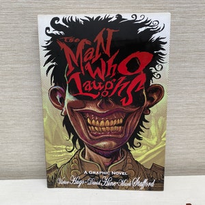 Könnte beinhalten: Das Cover von "The Man Who Laughs", einem Graphic Novel, zeigt eine verstörende Illustration eines lächelnden Gesichts. Der Titel ist in roter, stilisierter Schrift gehalten. Das Cover enthält den Text "A Graphic Novel" und die Namen der Autoren.