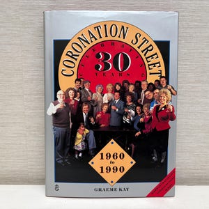May include: Book cover for "Coronation Street: 30 Years" with a cast photo. The cover is silver, with a circular logo reading "Coronation Street" and "30 Years". The years 1960 to 1990 are displayed in a diamond shape.