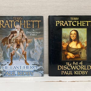 May include: Two hardcover books by Terry Pratchett, both illustrated by Paul Kidby. The first book is titled "The Last Hero: A Discworld Fable" and features a warrior with a sword. The second book is titled "The Art of Discworld" and features a woman with long, dark hair.