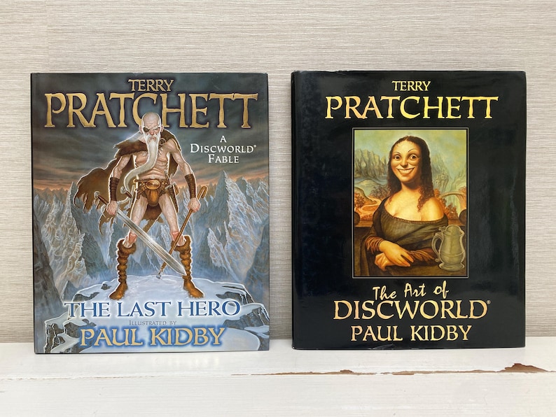 May include: Two hardcover books by Terry Pratchett, both illustrated by Paul Kidby. The first book is titled "The Last Hero: A Discworld Fable" and features a warrior with a sword. The second book is titled "The Art of Discworld" and features a woman with long, dark hair.