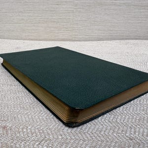 May include: A closed, hardcover book with a dark green cover and gold-colored page edges. The cover has a textured appearance. The book is lying flat on a light gray, textured surface.