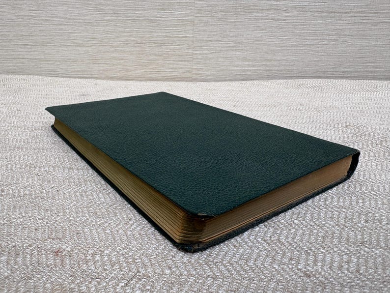 May include: A closed, hardcover book with a dark green cover and gold-colored page edges. The cover has a textured appearance. The book is lying flat on a light gray, textured surface.