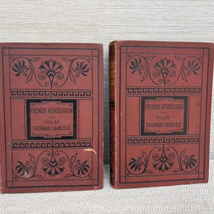 May include: Two antique books titled "French Revolution" by Thomas Carlyle. The books have a dark red cover with black decorative floral and geometric designs. The text is in black, and the books are displayed on a light grey surface.