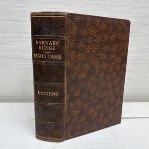 May include: A vintage hardcover book with a brown leather cover. The spine displays the titles "Barnaby Rudge" and "Edwin Drood" with the author's name, "Dickens," in gold lettering. The book has a textured appearance.