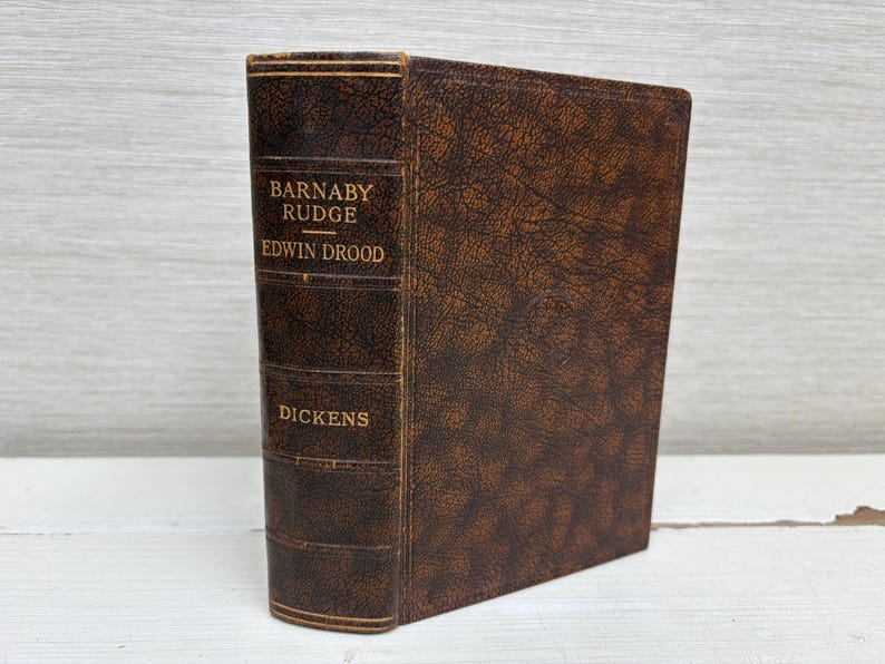 May include: A vintage hardcover book with a brown leather cover. The spine displays the titles "Barnaby Rudge" and "Edwin Drood" with the author's name, "Dickens," in gold lettering. The book has a textured appearance.