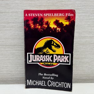 Könnte beinhalten: Ein Taschenbuch mit einem Cover, das eine Dinosaurier-Silhouette in einem gelben Kreis zeigt, mit dem Titel "JURASSIC PARK" in Weiß. Das Cover enthält auch den Text "A Steven Spielberg Film" und "The Bestselling Novel by MICHAEL CRICHTON".