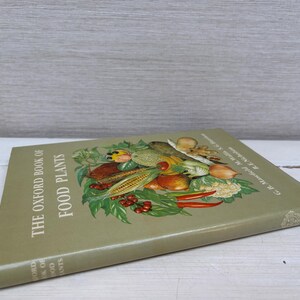 May include: A vintage book titled "The Oxford Book of Food Plants" with a green cover. The cover features a colorful illustration of various fruits and vegetables, including corn, tomatoes, and peppers. The book is resting on a white wooden surface.