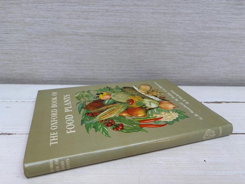 May include: A vintage book titled "The Oxford Book of Food Plants" with a green cover. The cover features a colorful illustration of various fruits and vegetables, including corn, tomatoes, and peppers. The book is resting on a white wooden surface.