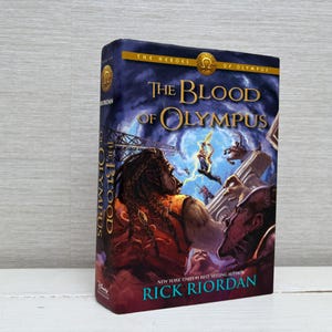 Puede incluir: Un libro de tapa dura titulado "The Blood of Olympus" de Rick Riordan. La portada presenta una ilustración dramática con figuras en acción, sobre un fondo azul oscuro y dorado. El libro forma parte de la serie "The Heroes of Olympus".