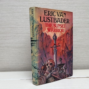 Puede incluir: Un libro de bolsillo vintage titulado "The Sunset Warrior" de Eric Van Lustbader. La portada presenta una ilustración de fantasía con una espada, una calavera y un fondo de puesta de sol ardiente. El lomo muestra el nombre del autor y el título.