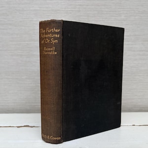 May include: A vintage hardcover book titled "The Further Adventures of Dr. Syn" by Russell Thorndike. The book has a dark brown spine with gold lettering and a black cover. Published by Rich & Cowan.