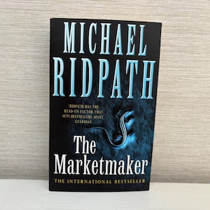 May include: A hardcover book titled "The Marketmaker" by Michael Ridpath. The cover is predominantly black with the author's name in large, light blue letters. The title is in white, and a blue and black illustration is featured.