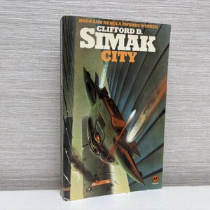 May include: A book cover with a futuristic illustration of a spaceship flying over a city. The title of the book is "SIMAK CITY" by Clifford D. Simak, a Hugo and Nebula Award winner.