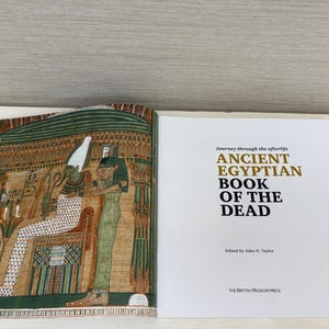 May include: An open book with colorful illustrations of ancient Egyptian figures and hieroglyphs. The right page displays the title "ANCIENT EGYPTIAN BOOK OF THE DEAD" in gold and black text. Edited by John H. Taylor, published by The British Museum Press.