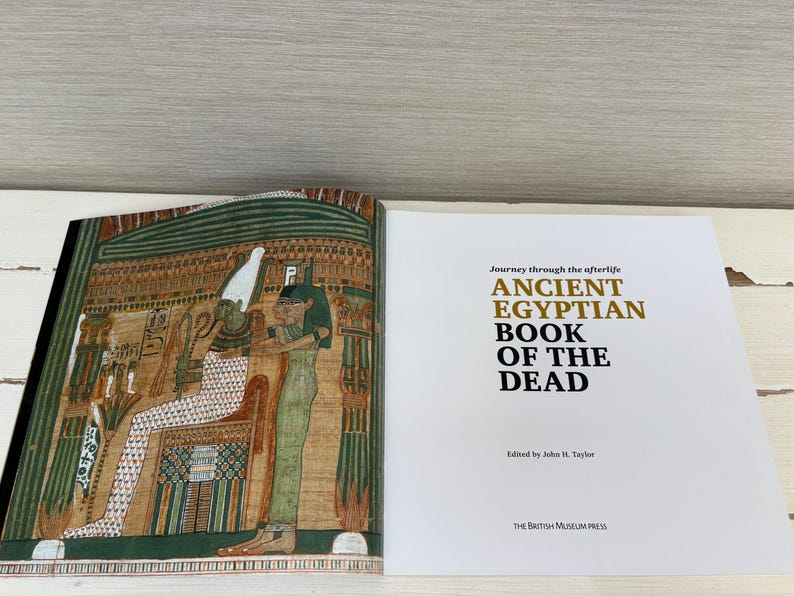 May include: An open book with colorful illustrations of ancient Egyptian figures and hieroglyphs. The right page displays the title "ANCIENT EGYPTIAN BOOK OF THE DEAD" in gold and black text. Edited by John H. Taylor, published by The British Museum Press.