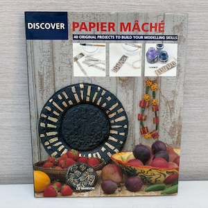 Peut inclure: Un livre intitulé "Discover Papier Mâché" avec une image colorée de fruits et une assiette sur la couverture. Le livre propose 40 projets originaux pour développer vos compétences en modelage.