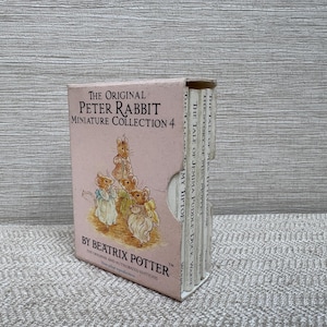 Peut inclure: Un coffret de livres miniatures intitulé "The Original Peter Rabbit Miniature Collection 4" par Beatrix Potter. La boîte est rose clair avec des illustrations de personnages de Pierre Lapin. L'ensemble comprend plusieurs petits livres.