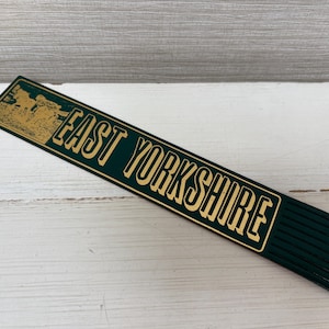 May include: A dark green wooden ruler with the words "EAST YORKSHIRE" in gold lettering. The ruler has a gold illustration on the left side. The ruler is lying on a white wooden surface.