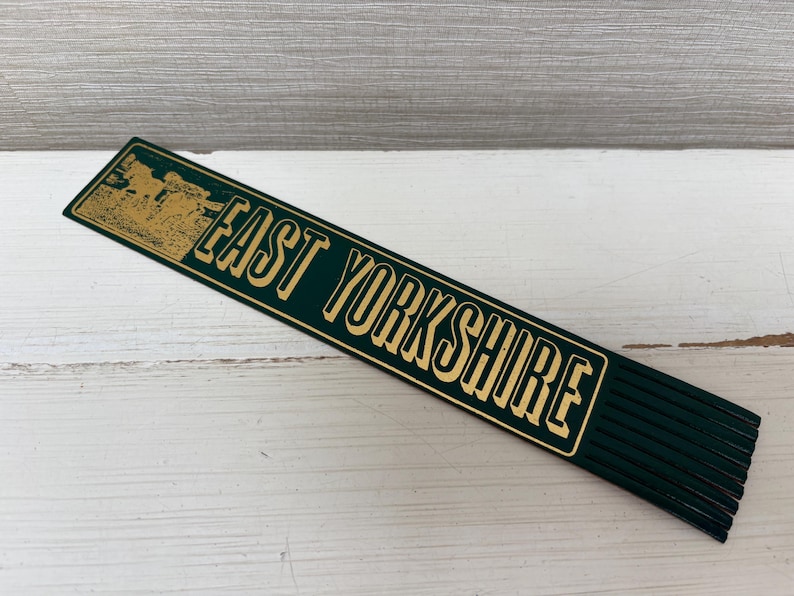 May include: A dark green wooden ruler with the words "EAST YORKSHIRE" in gold lettering. The ruler has a gold illustration on the left side. The ruler is lying on a white wooden surface.