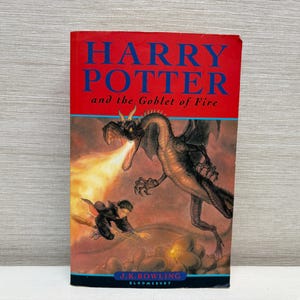 Può includere: Un libro con copertina rigida intitolato Harry Potter and the Goblet of Fire di J.K. Rowling. La copertina rossa presenta un grande drago alato scuro che sputa fuoco verso una piccola figura che vola su una scopa. Il titolo è stampato in blu.