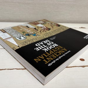 May include: A hardcover book titled "Ancient Egyptian Book of the Dead" with a black cover and gold lettering. The book's cover features illustrations of ancient Egyptian figures and hieroglyphs. The book is open, revealing the pages.