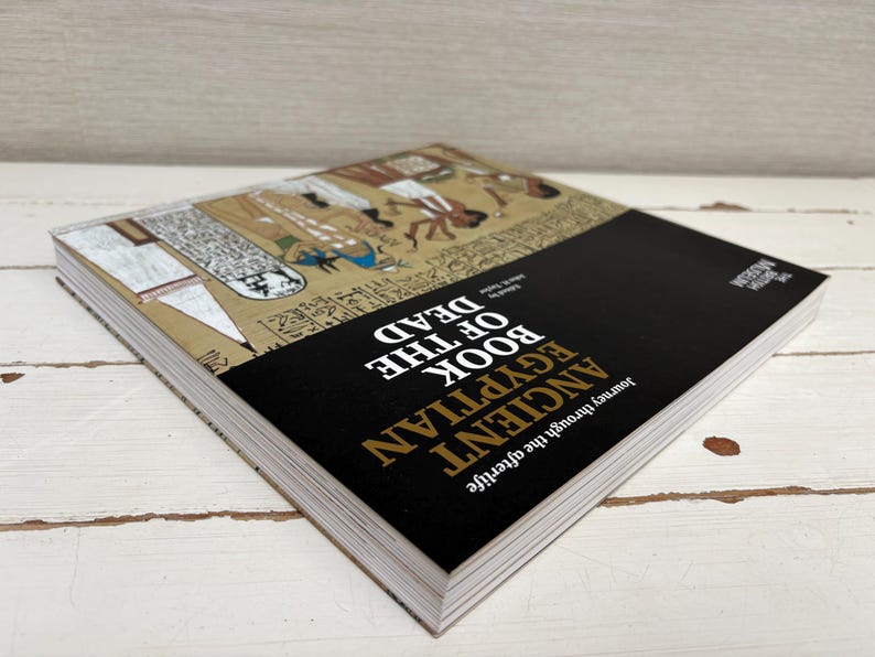 May include: A hardcover book titled "Ancient Egyptian Book of the Dead" with a black cover and gold lettering. The book's cover features illustrations of ancient Egyptian figures and hieroglyphs. The book is open, revealing the pages.