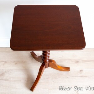 May include: A dark brown wooden side table with a square top and a twisted, turned, wooden pedestal base. The table is on a light wood floor.